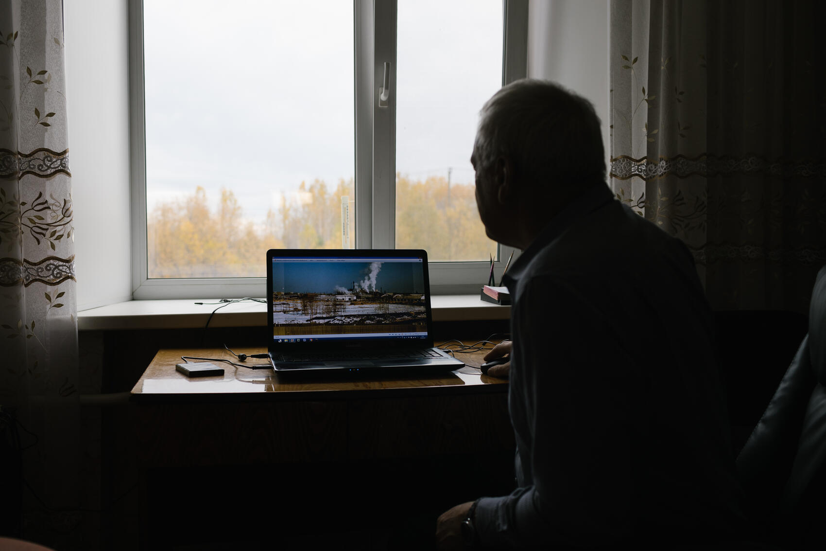 Владимир рассказывает о прошлом Ивделя. На компьютере изображение Лесозавода в прошлом. За окном осенний пейзаж —- то, что от осталось от закрывшегося Лесозавода